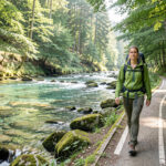 Une randonneuse marche sur un sentier balisé longeant une rivière claire au cœur d’une forêt lumineuse et naturelle.