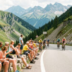 Des hommes et des femmes spectateurs installés au bord d’une route sinueuse des Alpes, attendant le passage d’une course cycliste près d’Orcières-Merlette sous une lumière estivale.