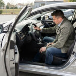 Un homme examine attentivement l’intérieur d’une voiture moderne d’occasion garée sur un parking éclairé par la lumière naturelle.
