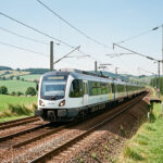 Train régional moderne à batteries circulant sur une voie de campagne et quittant une zone équipée de caténaires, sous une lumière naturelle et lumineuse.