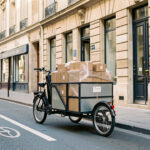 Triporteur électrique professionnel chargé de colis, stationné près d’une piste cyclable devant des commerces aux façades en pierre claire du centre-ville de Bordeaux.